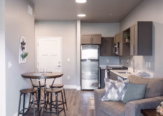 Interior view of a modern senior living apartment at The Pointe at Meridian featuring a small dining area with a round table and four stools, a kitchen with stainless steel refrigerator, microwave, and sink, and a cozy living area with a gray couch and decorative pillows.