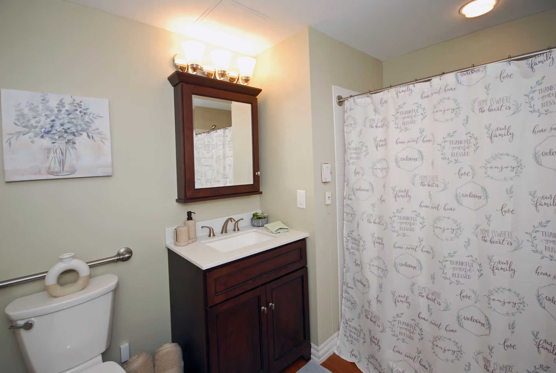 A bathroom with a white toilet, a dark wood vanity with a white countertop and sink, a wall-mounted mirror with lights above it, a shower with a curtain featuring various inspirational words and phrases, and a decorative painting of a vase with flowers on the wall.