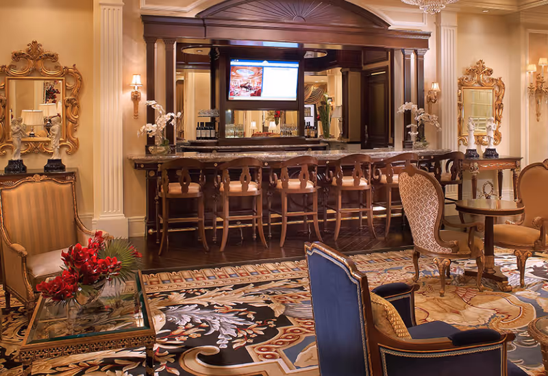 Elegant interior lounge area with a long wooden bar and several bar stools. The room features ornate furniture including upholstered chairs and a glass coffee table with a red floral arrangement. The walls are decorated with mirrors, sconces, and classical columns, and a large patterned rug covers the floor.