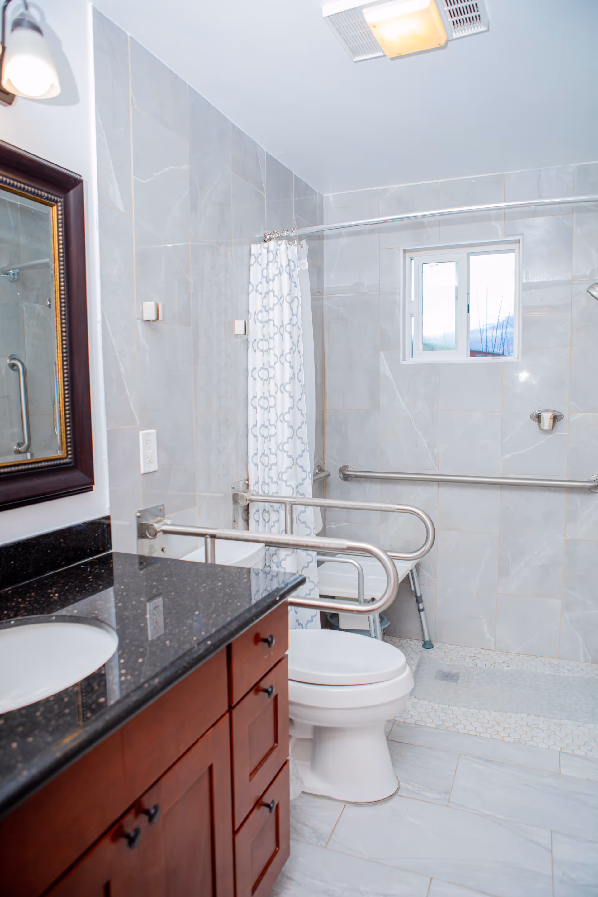 A modern bathroom with gray tiled walls and floor, featuring a white toilet with stainless steel safety rails on both sides, a shower area with a patterned white and gray shower curtain, a small window, and a wooden vanity with a black granite countertop and a sink. There is also a mirror with a dark wooden frame and a light fixture above it.