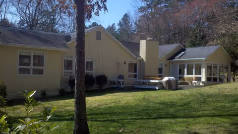 Single-story beige senior living facility with a wooden deck, chairs, and grassy lawn.