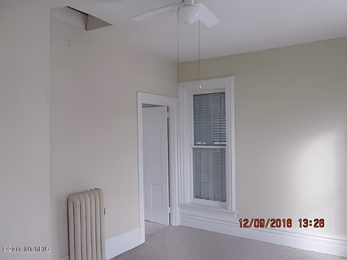 Empty room with beige walls, a white ceiling fan, a radiator, a window with blinds, and an open door leading to another room. The ceiling has a damaged area with a hole.