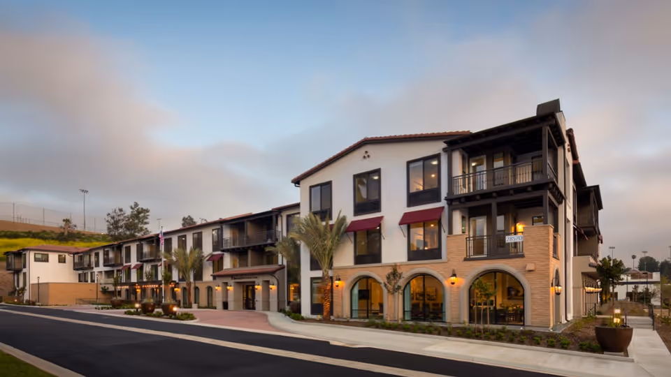 Exterior view of a modern three-story senior living facility building with white walls, large windows, balconies, and red awnings, surrounded by palm trees and landscaping under a cloudy sky.