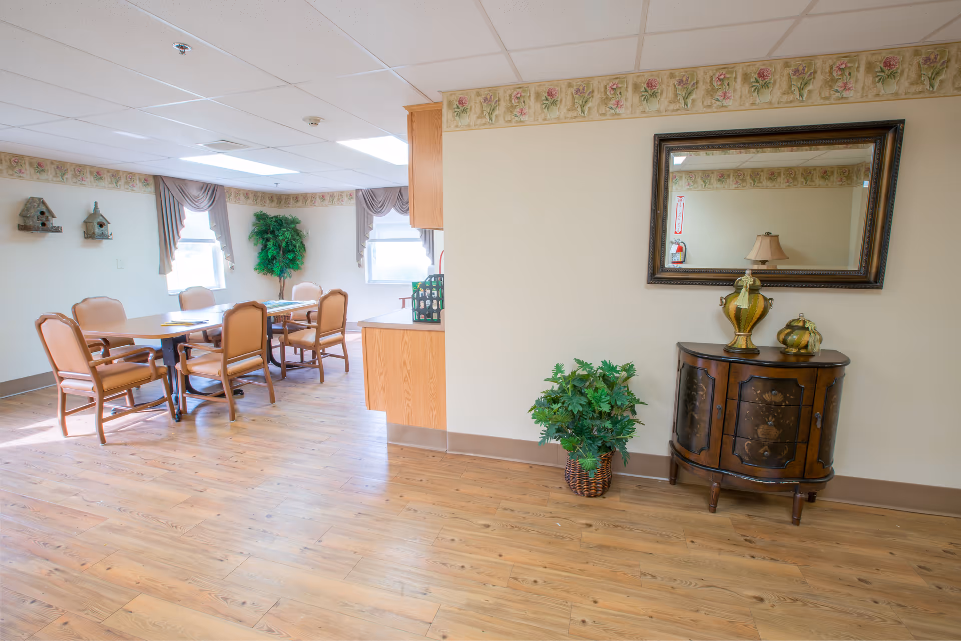 A bright and clean dining area in a senior living facility with a wooden table surrounded by six cushioned chairs. The room features light-colored walls with floral wallpaper border near the ceiling, two windows with draped curtains, a potted plant in the corner, and a small wooden cabinet with decorative vases and a mirror above it.