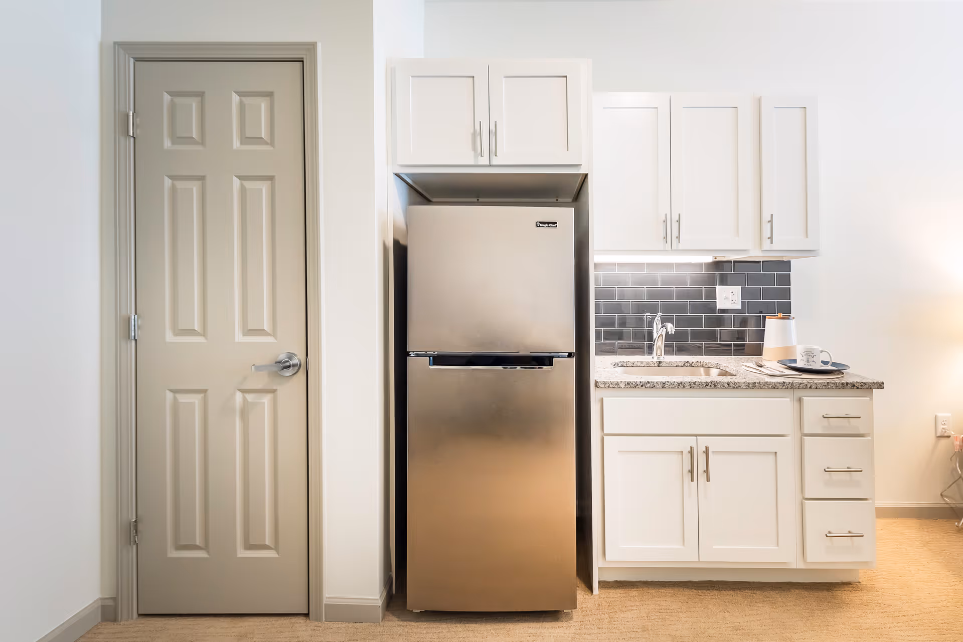 A compact kitchen area featuring a stainless steel refrigerator, white cabinets with silver handles, a granite countertop with a sink, and a dark gray tiled backsplash. To the left of the refrigerator is a closed beige door, and to the right on the countertop are a cup, a small container, and a plate with utensils.