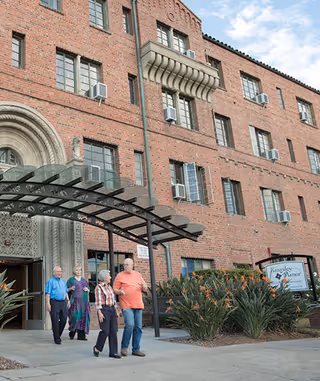 Four elderly people walking outside the entrance of a large brick building with multiple windows and air conditioning units. The building has an arched doorway with decorative stonework and a black metal canopy over the entrance. There are plants and flowers near the entrance, and a sign that reads 'Kingsley Manor' is visible on the right side.
