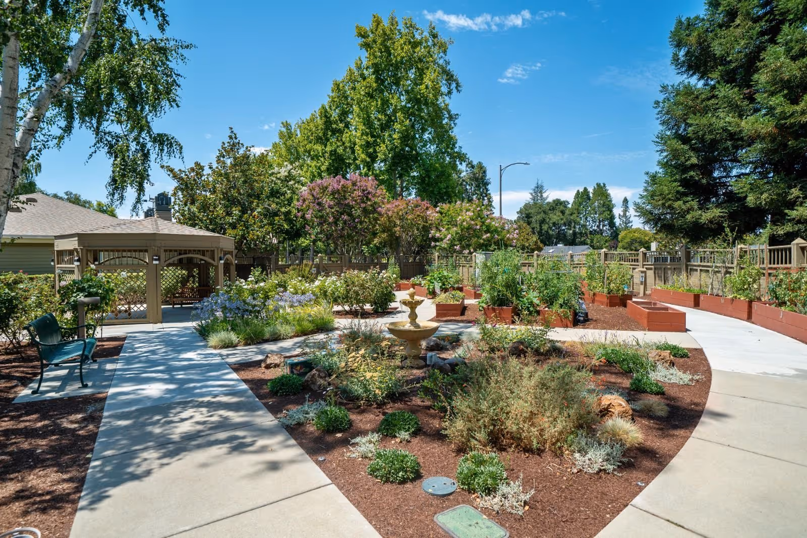 A sunny outdoor garden area with a paved walkway, green benches, a small fountain, various plants and shrubs, raised garden beds, and a gazebo. Trees and a wooden fence surround the garden under a clear blue sky.