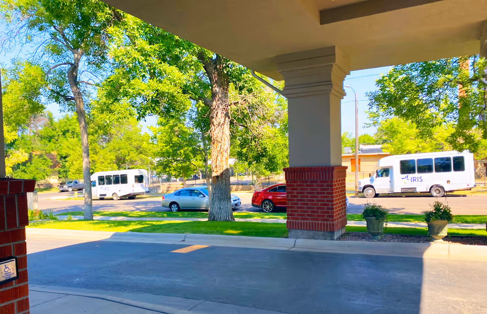 View from under a covered entrance showing a driveway with a brick column and two potted plants. Across the street, there are two white shuttle vans with the word 'IRIS' on them, parked near green trees and a few cars.