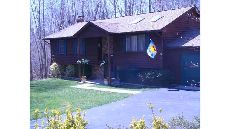 Exterior view of a single-story brown wooden house with a sloped roof, blue shutters, a small front porch, and a driveway leading to a garage. There is a colorful flag hanging near the front entrance, and leafless trees are visible in the background.