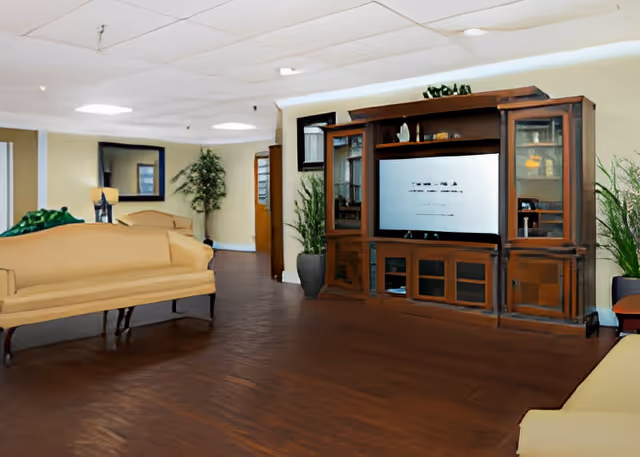 Interior view of a senior living facility common area with wooden flooring, beige sofas, a large wooden entertainment center with a television, potted plants, and a mirror on the wall.