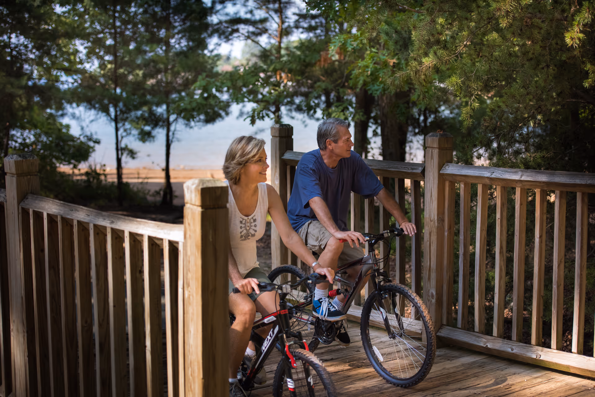 A man and a woman riding bicycles on a wooden bridge surrounded by trees with a lake visible in the background.
