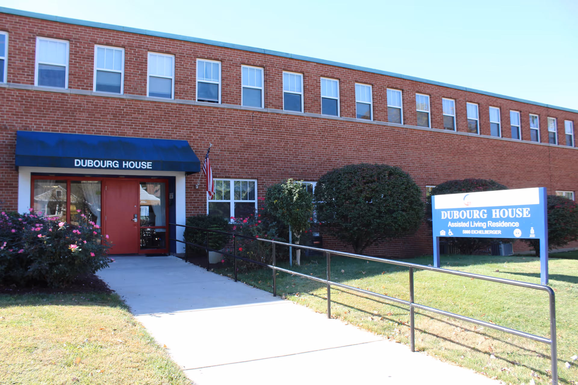 Exterior view of Dubourg House, a two-story brick building with multiple windows. The entrance has a blue awning with the name 'Dubourg House' above red double doors. There is a ramp with handrails leading to the entrance. A blue sign on the lawn reads 'Dubourg House Assisted Living Residence' along with the address 5800 Bechelberger. Bushes and a small tree are planted near the building, and an American flag is mounted near the entrance.