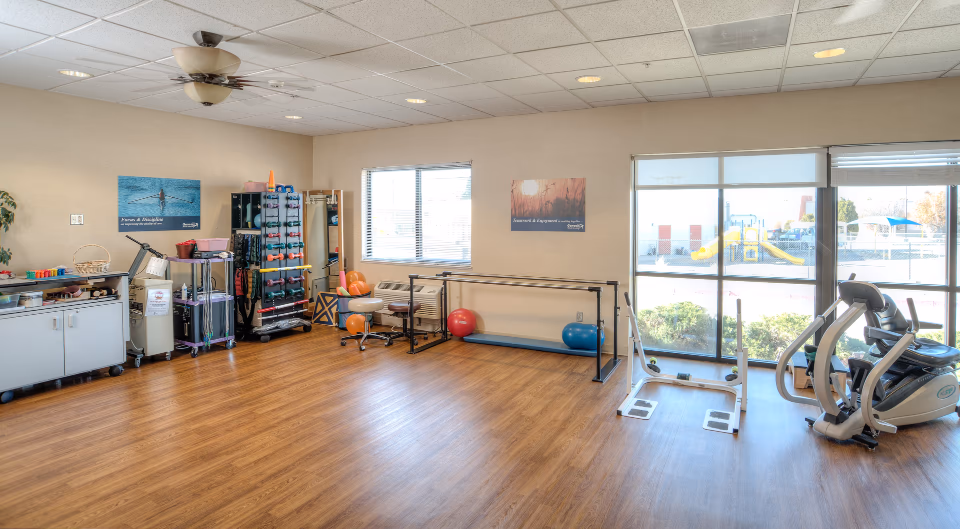 A spacious rehabilitation exercise room with wooden flooring, various exercise equipment including weights, exercise balls, parallel bars, and a stationary bike. Large windows allow natural light to fill the room, and there are motivational posters on the walls.