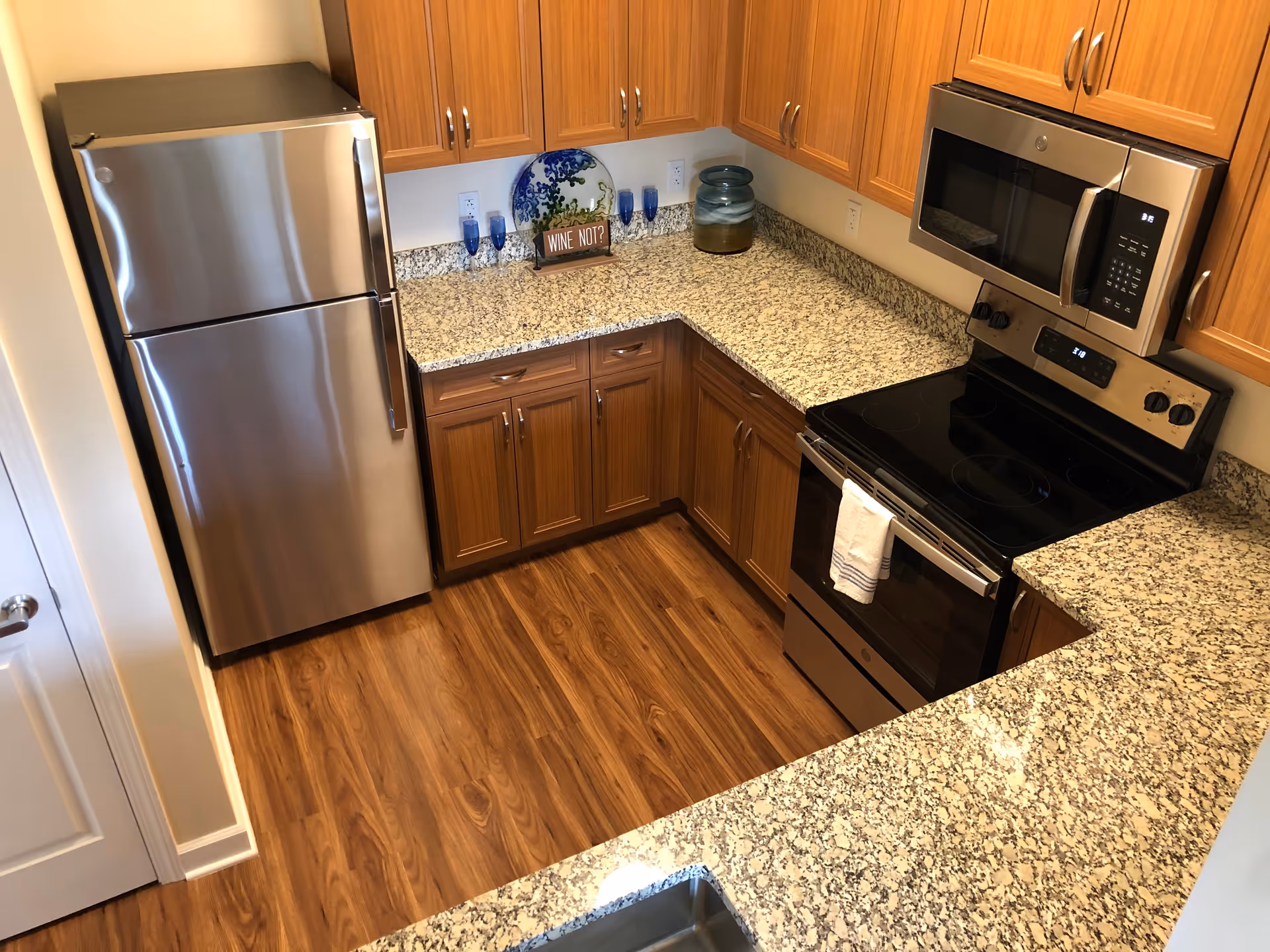 Small modern kitchen with stainless steel refrigerator and stove, granite countertops, wooden cabinets, and wood-look flooring.
