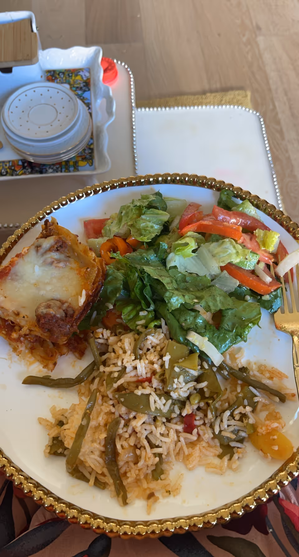 A plate with a serving of lasagna, a fresh salad with lettuce and tomatoes, and a portion of rice mixed with green beans and other vegetables, placed on a table with a decorative gold-rimmed edge. In the background, there is a white tray holding a stack of small plates and a napkin holder.