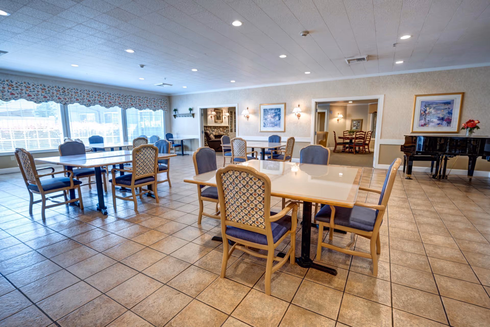 Spacious senior dining room with several tables and upholstered chairs, large windows, tiled floor, and a piano against the wall.