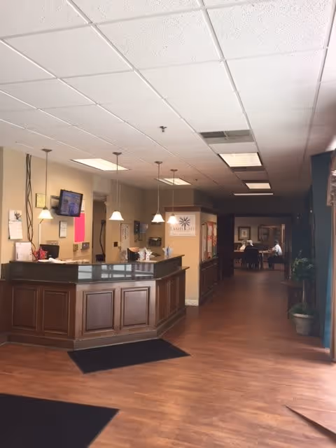 Interior view of a senior living facility reception area with a wooden front desk, hanging pendant lights, a wall-mounted TV, and a hallway leading to a room where two people are seated at a table. The floor is wooden, and there are plants and framed pictures along the walls.