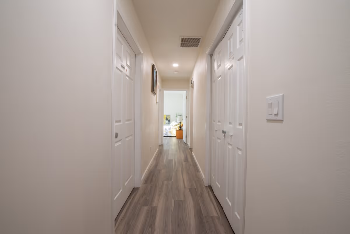 A narrow hallway with light-colored walls and wood flooring. There are multiple white doors on both sides of the hallway, and a room with a bed and some furniture is visible at the end of the hallway. The ceiling has recessed lighting and an air vent.