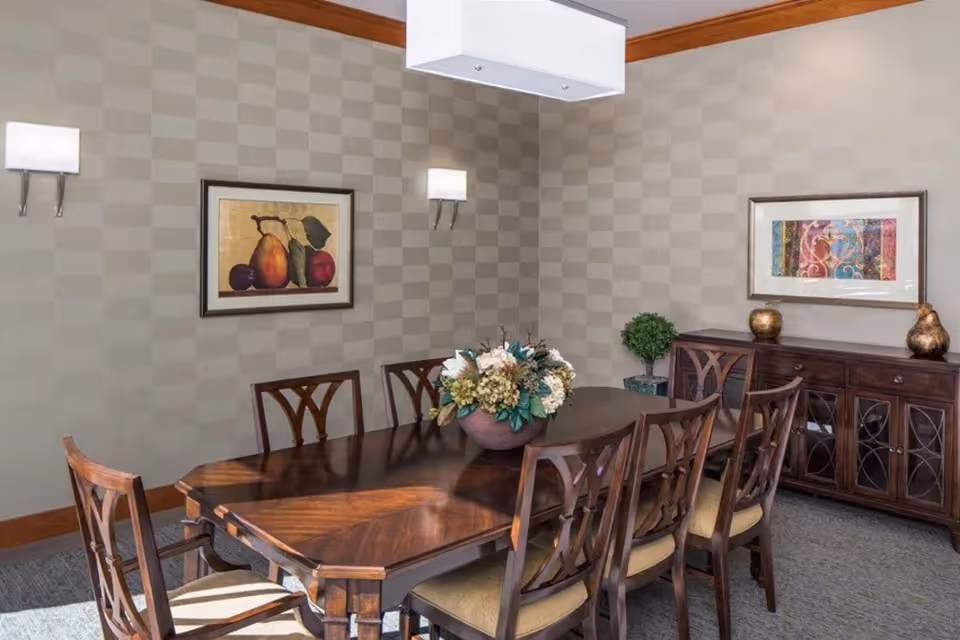 A dining room with a dark wooden table surrounded by eight matching chairs with cushioned seats. A floral centerpiece is placed on the table. The walls have a checkered pattern wallpaper and are decorated with two framed artworks. There is a wooden sideboard with decorative items and a small potted plant. The room is lit by wall sconces and a rectangular ceiling light fixture.