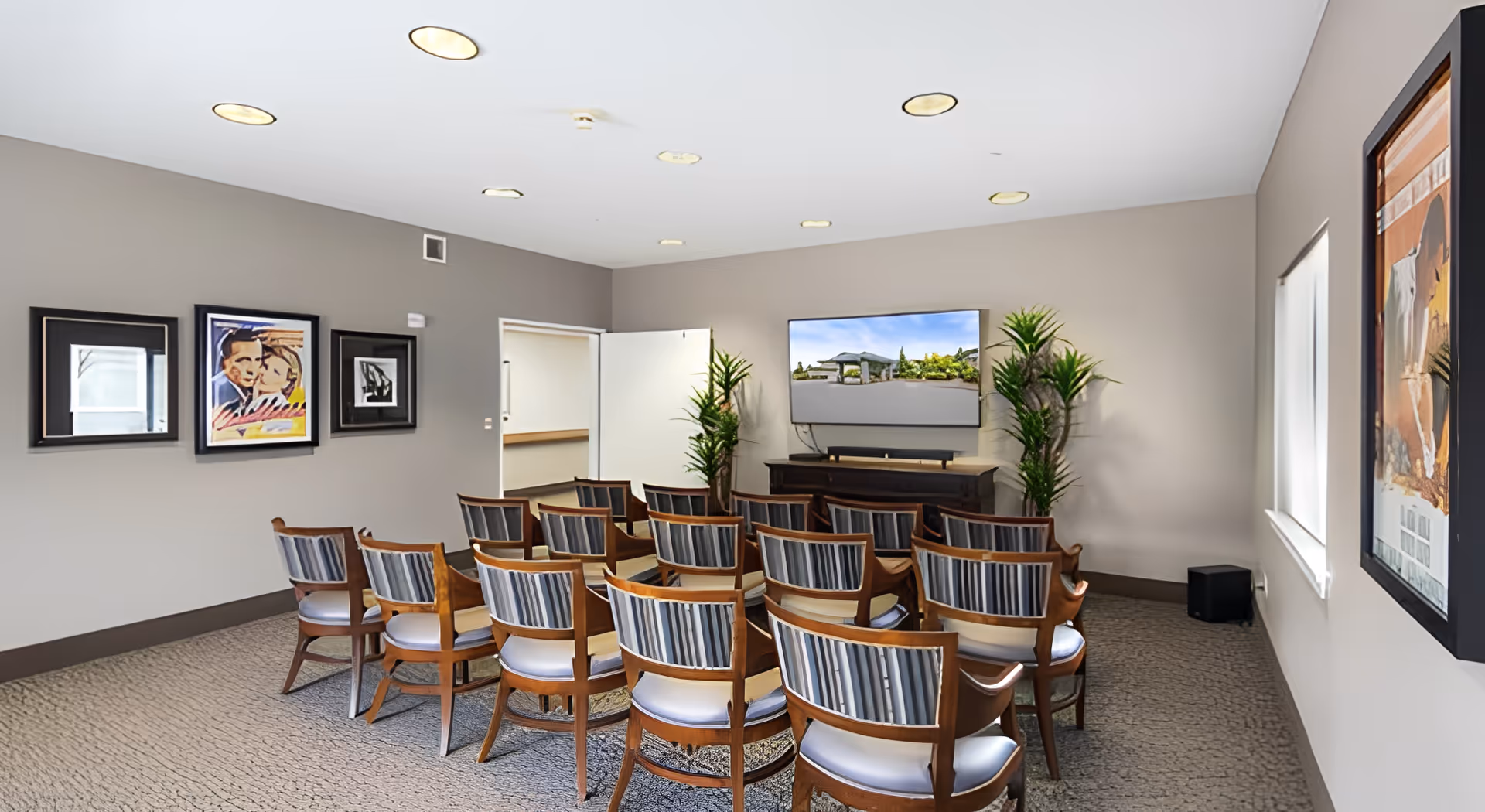 A small conference or media room with rows of wooden chairs with striped cushions facing a flat-screen TV mounted on the wall. The room has beige walls, carpeted floor, framed artwork on the walls, two potted plants flanking the TV, and recessed ceiling lights.