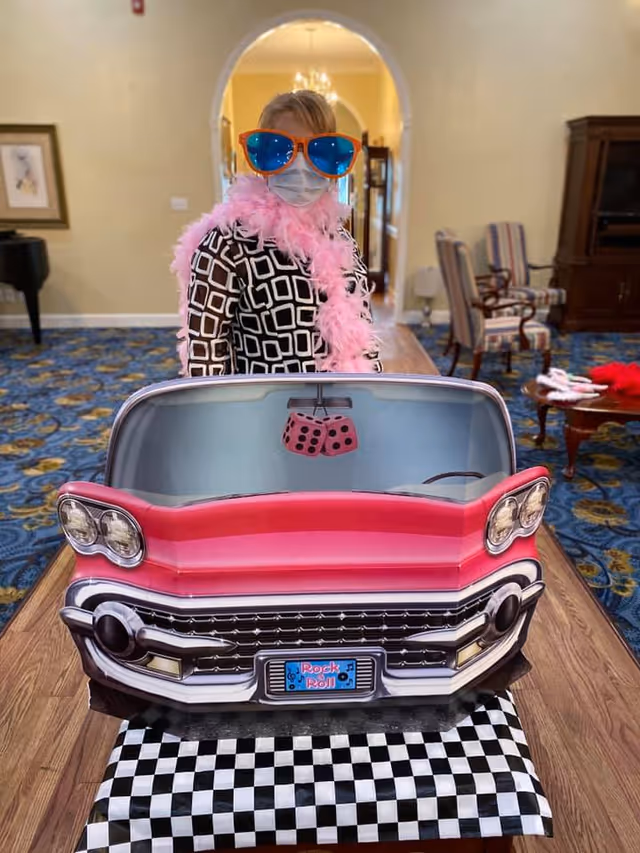 A person wearing oversized blue sunglasses, a pink feather boa, and a face mask stands behind a cardboard cutout of a red vintage car with a 'Rock & Roll' license plate, set indoors in a room with patterned blue carpet and striped chairs.