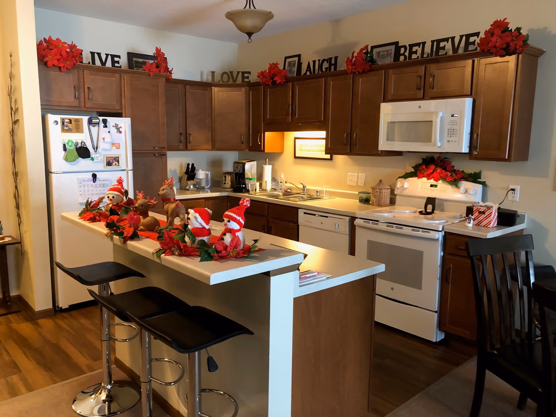 A cozy kitchen with wooden cabinets, white appliances including a refrigerator, stove, microwave, and dishwasher. The kitchen island has three black bar stools and is decorated with Christmas-themed figurines and red poinsettias. Above the cabinets are decorative words 'LIVE', 'LOVE', 'LAUGH', and 'BELIEVE' along with more red poinsettias. The floor is wooden and there is a chair partially visible on the right side.