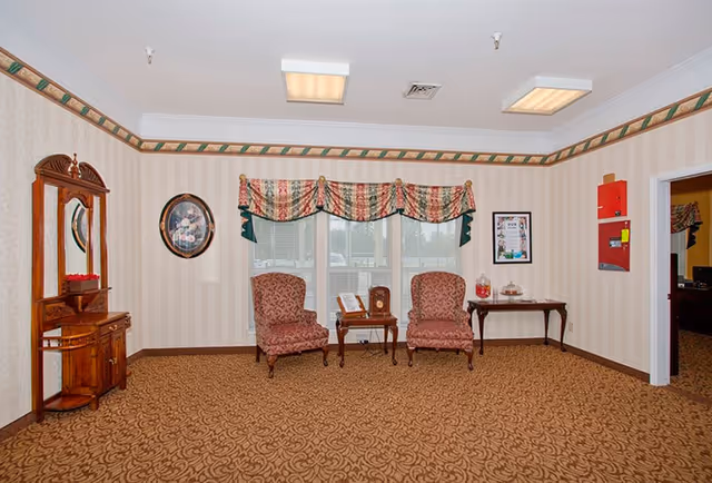 A sitting area in a senior living facility with two patterned armchairs placed in front of three windows with valance curtains. Between the chairs is a small wooden table with a clock and a book. To the left is a wooden cabinet with a mirror and decorative items. On the right wall, there is a framed picture and a red fire safety panel. The room has patterned carpet and wallpaper with a decorative border near the ceiling.