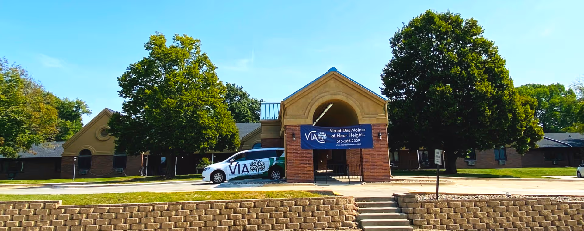 Brick single-story senior living facility front with a covered entrance, Via-branded van, sign, and trees.