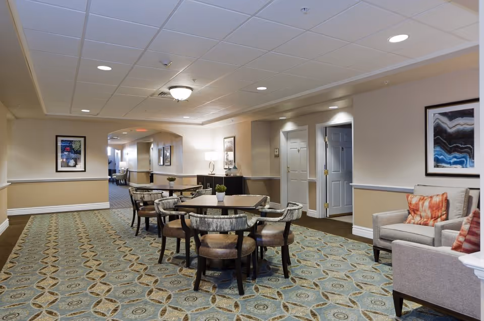 Interior view of a senior living facility common area with patterned carpet, several tables with chairs, a beige armchair with orange cushions, framed artwork on the walls, and soft lighting from ceiling fixtures and a table lamp.