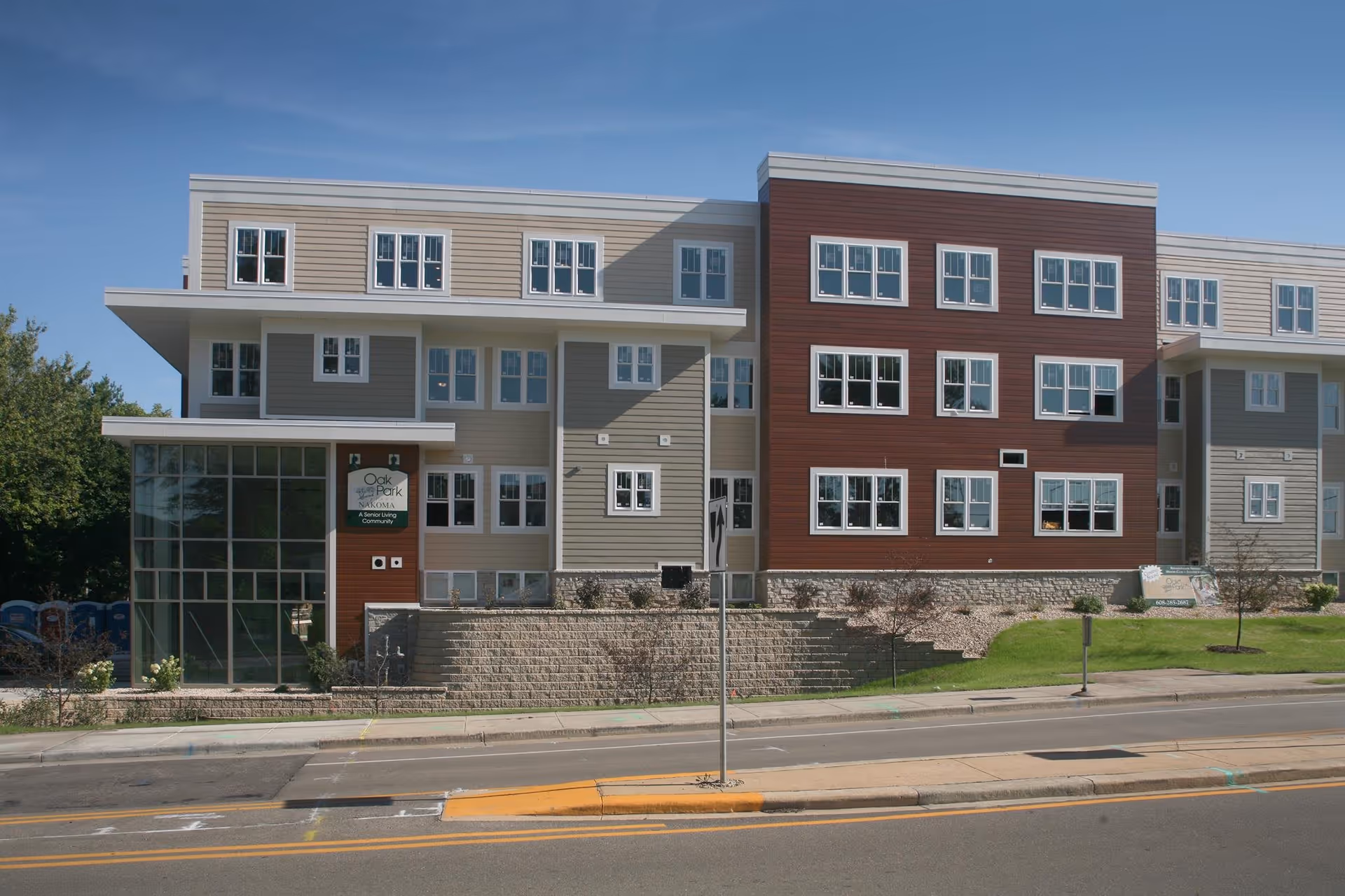Exterior view of Oak Park Place Nakoma, a senior living community building with multiple windows, beige and brown siding, and a glass entrance on the left side. The building is situated along a street with a sidewalk and some landscaping in front.