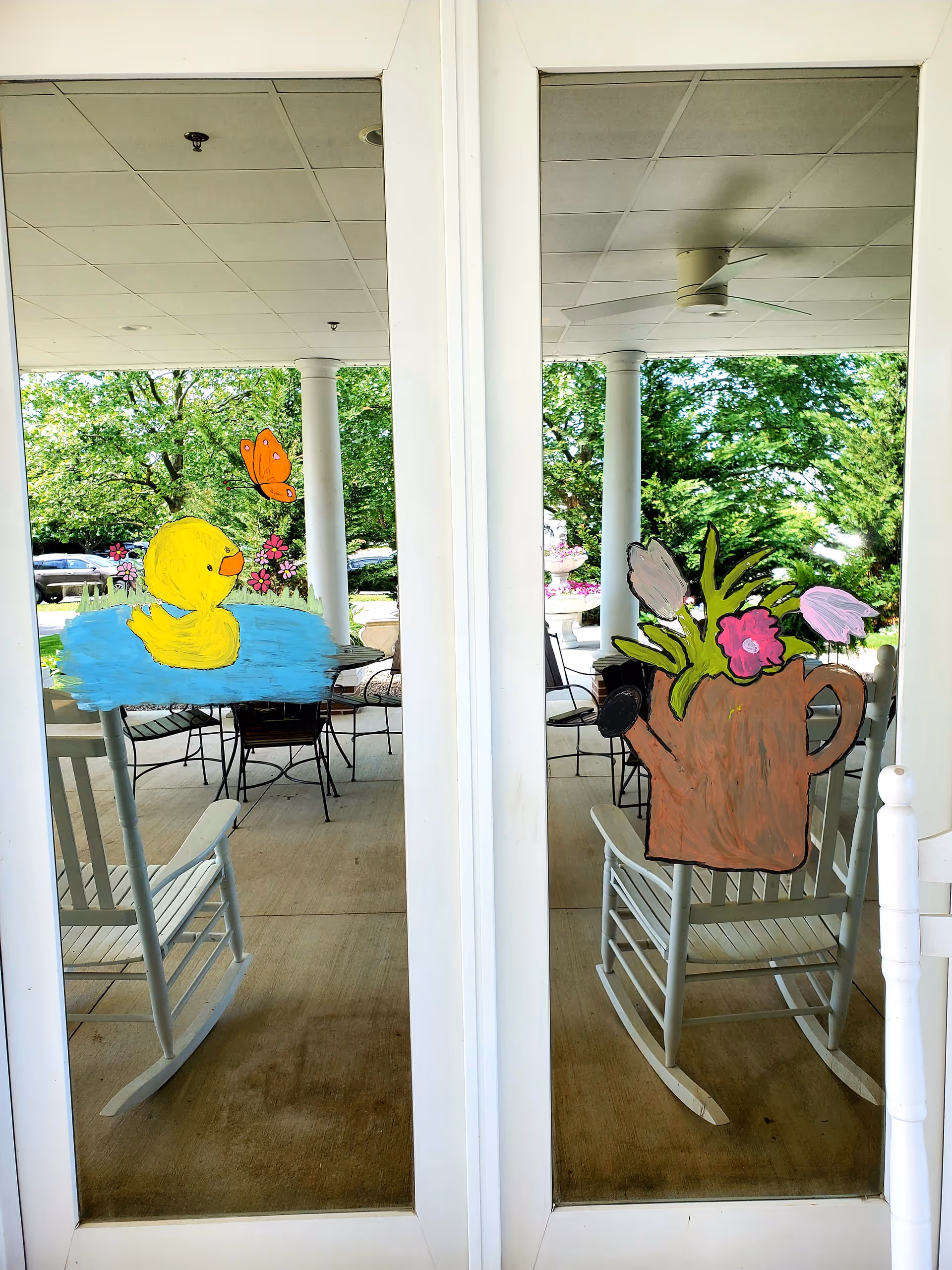 Glass doors with painted decorations of a yellow rubber duck in blue water with a butterfly and flowers on the left door, and a brown watering can with pink flowers on the right door. Behind the doors is a covered outdoor patio area with white rocking chairs, metal chairs, tables, white columns, and green trees in the background.