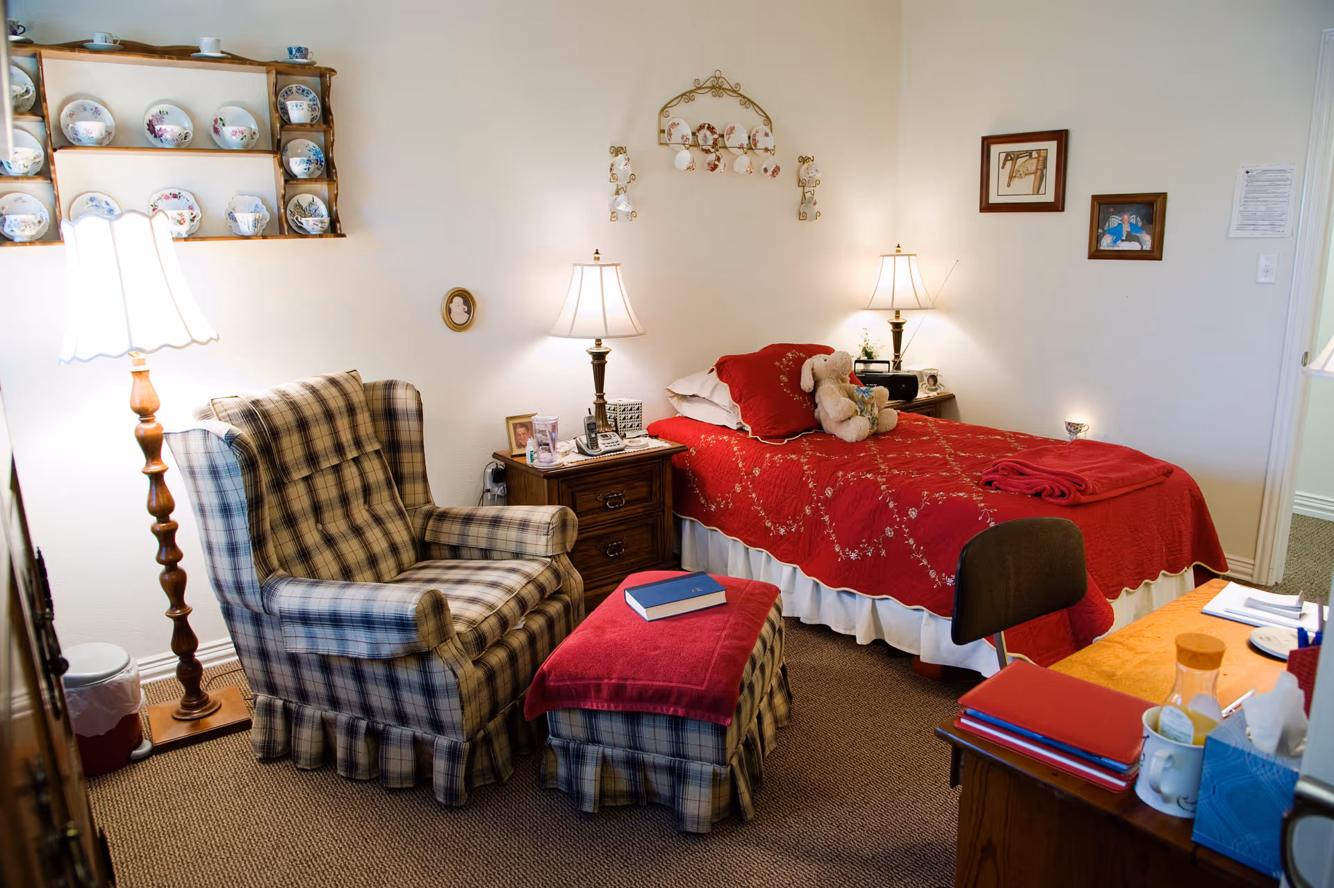 A cozy bedroom in an assisted living facility featuring a single bed with a red quilt and a teddy bear on it. There is a plaid armchair with a matching ottoman covered by a red cloth, a wooden nightstand with a lamp, framed pictures on the wall, and a wooden shelf displaying decorative plates. A floor lamp stands beside the armchair, and a desk with various items is partially visible in the foreground.