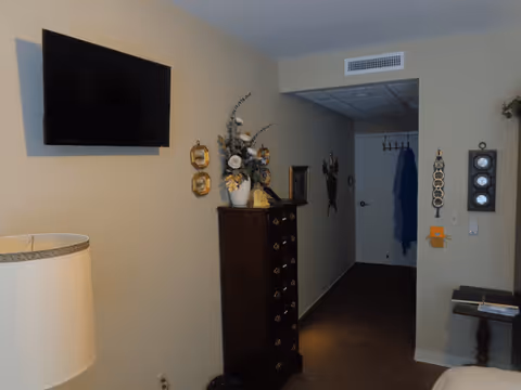 Bedroom interior with a wall-mounted TV, tall dresser topped with a floral arrangement, lamp, and a hallway leading to a door.
