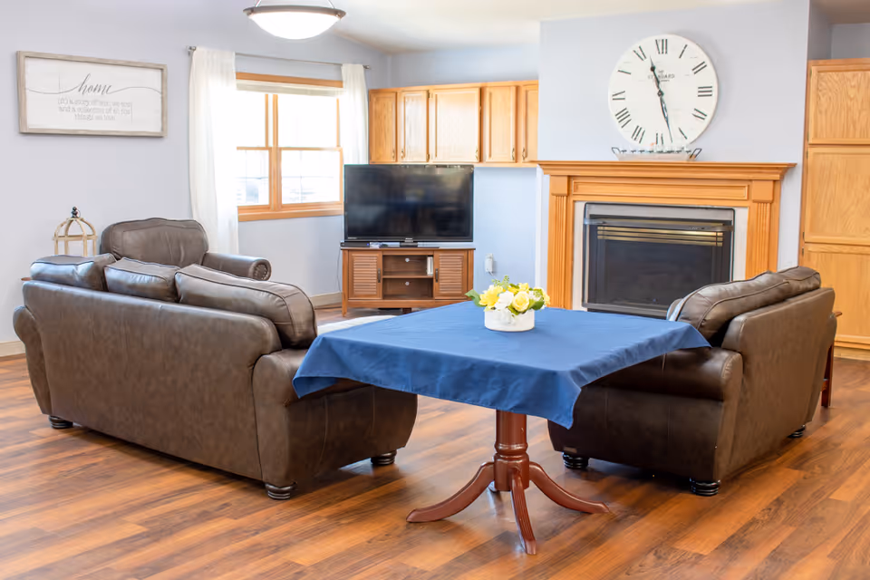 Cozy sitting area with two brown leather sofas facing a TV and fireplace and a small table with a blue cloth and flowers in the center.