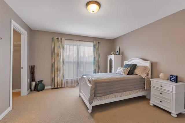 A neatly arranged bedroom with a white wooden bed featuring gray bedding and decorative pillows. There is a white nightstand with drawers on the right side of the bed, holding a small lamp and a framed sign. A white cabinet is positioned against the wall near the window, which is covered with sheer white curtains and patterned drapes. The walls are painted beige, and the floor is carpeted in a light brown color. Two decorative vases are placed on the floor near the doorway on the left.