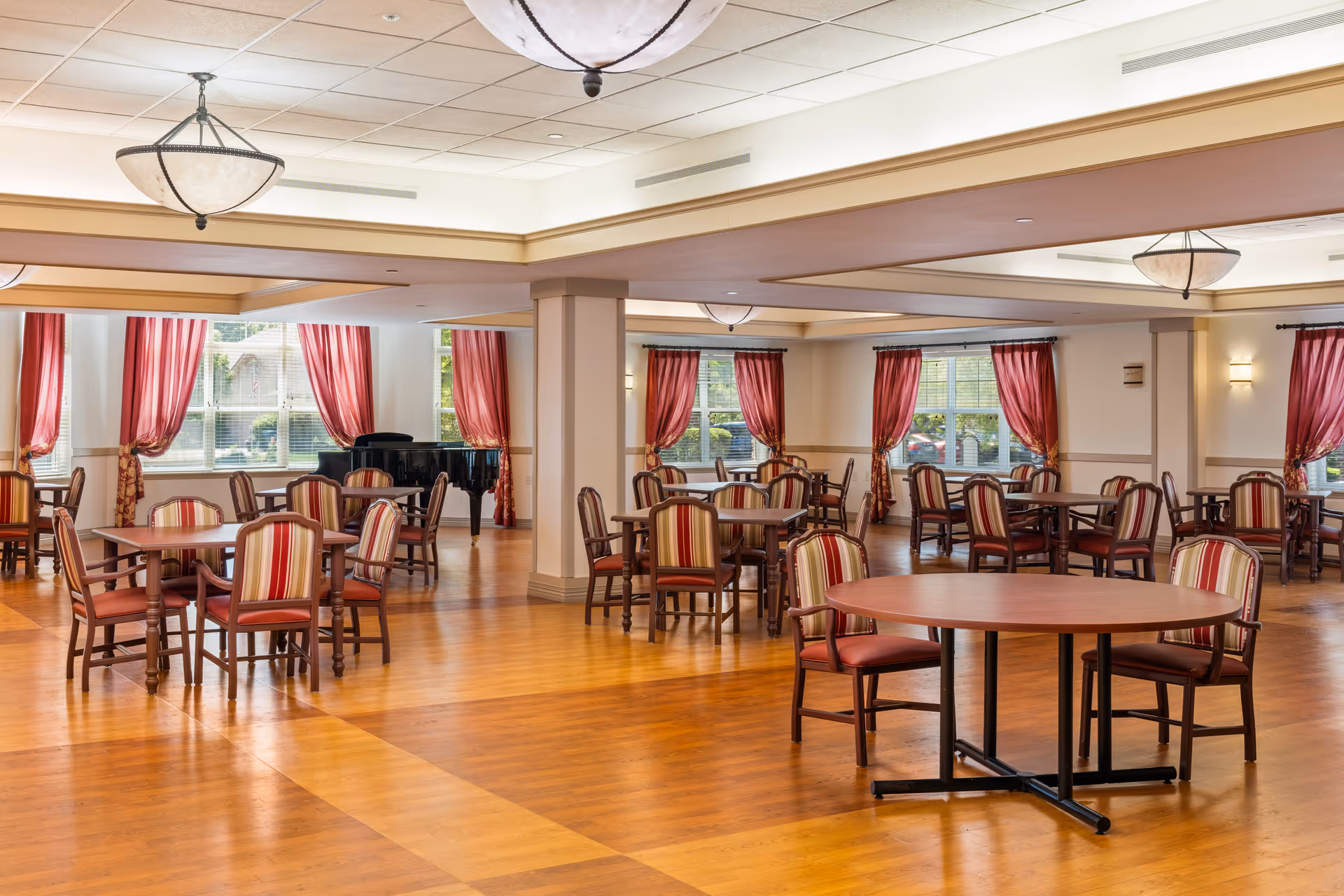 Large dining/activity room with multiple round and rectangular tables, striped upholstered chairs, red curtains on windows, and a black grand piano.