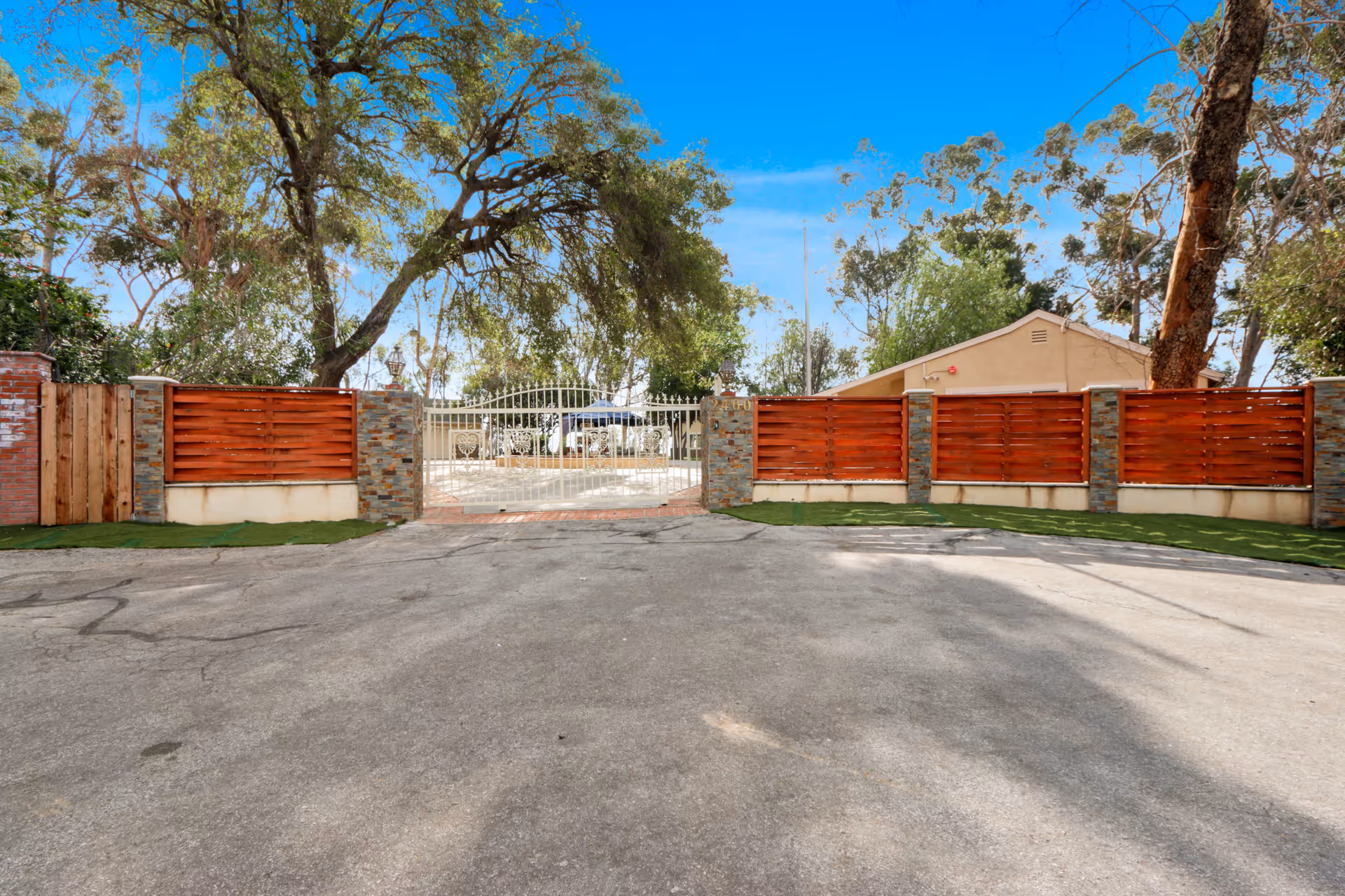 View of a gated entrance to a property with a white metal gate and stone pillars, surrounded by wooden fencing and large trees under a clear blue sky.