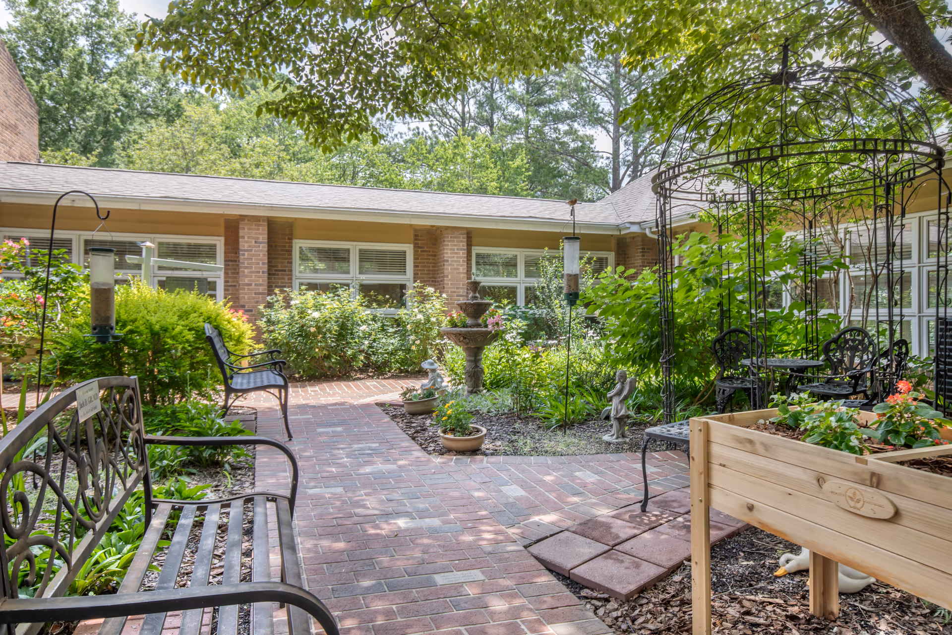 A peaceful outdoor courtyard garden at Brookdale Chambrel Roswell featuring brick pathways, metal benches, a tiered water fountain, bird feeders, lush greenery, flowering plants, a wooden planter box, and a decorative metal gazebo with chairs and a table underneath.
