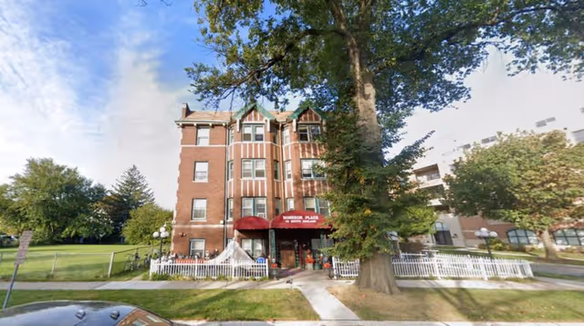 Exterior view of a three-story brick building with a red canopy entrance labeled 'Windsor Place'. The building is surrounded by a white picket fence, trees, and a grassy lawn under a partly cloudy sky.