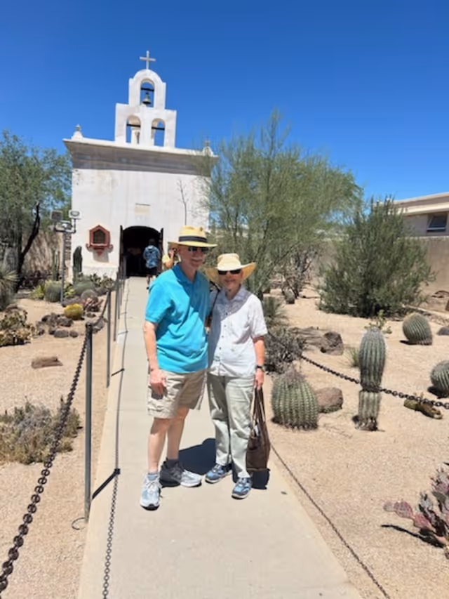 An elderly couple wearing hats and sunglasses stands on a paved walkway in front of a small white chapel with a bell tower and cross. The surrounding area is a desert landscape with various cacti and desert plants under a clear blue sky.