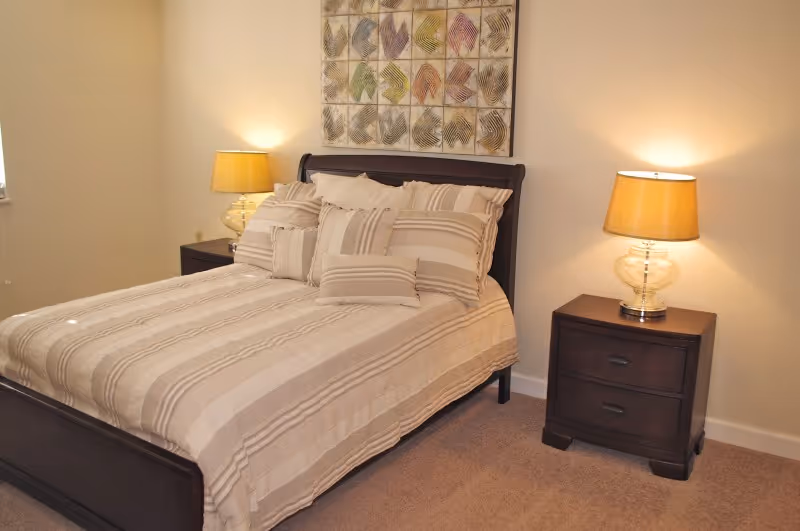 A neatly made bed with beige and white striped bedding and multiple pillows in a bedroom. There are two dark wooden nightstands on either side of the bed, each with a lamp that has a beige lampshade. Above the bed is a decorative wall art piece featuring a grid of colorful leaf patterns.