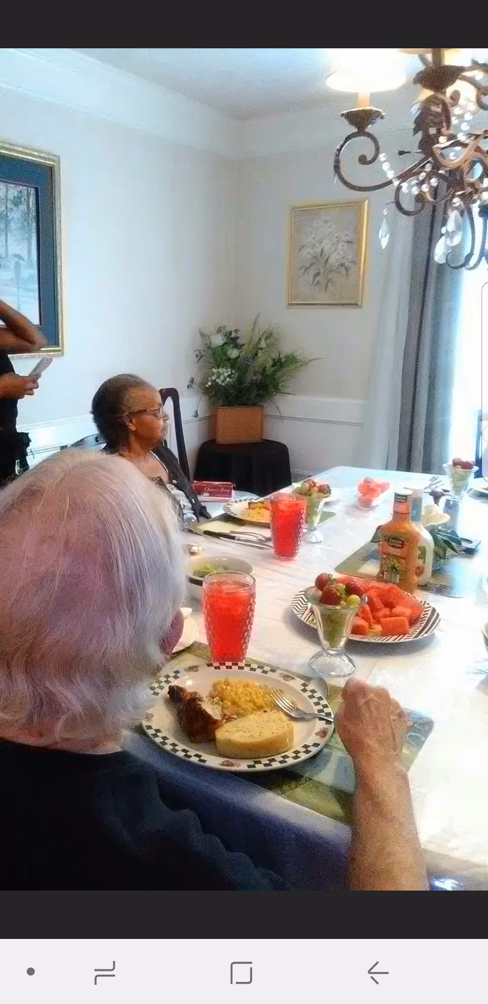 Two elderly women seated at a dining table set with plates of food, glasses of red drink, and bowls of fruit. The room has light-colored walls with framed artwork, a chandelier, and a floral arrangement on a small table in the corner.