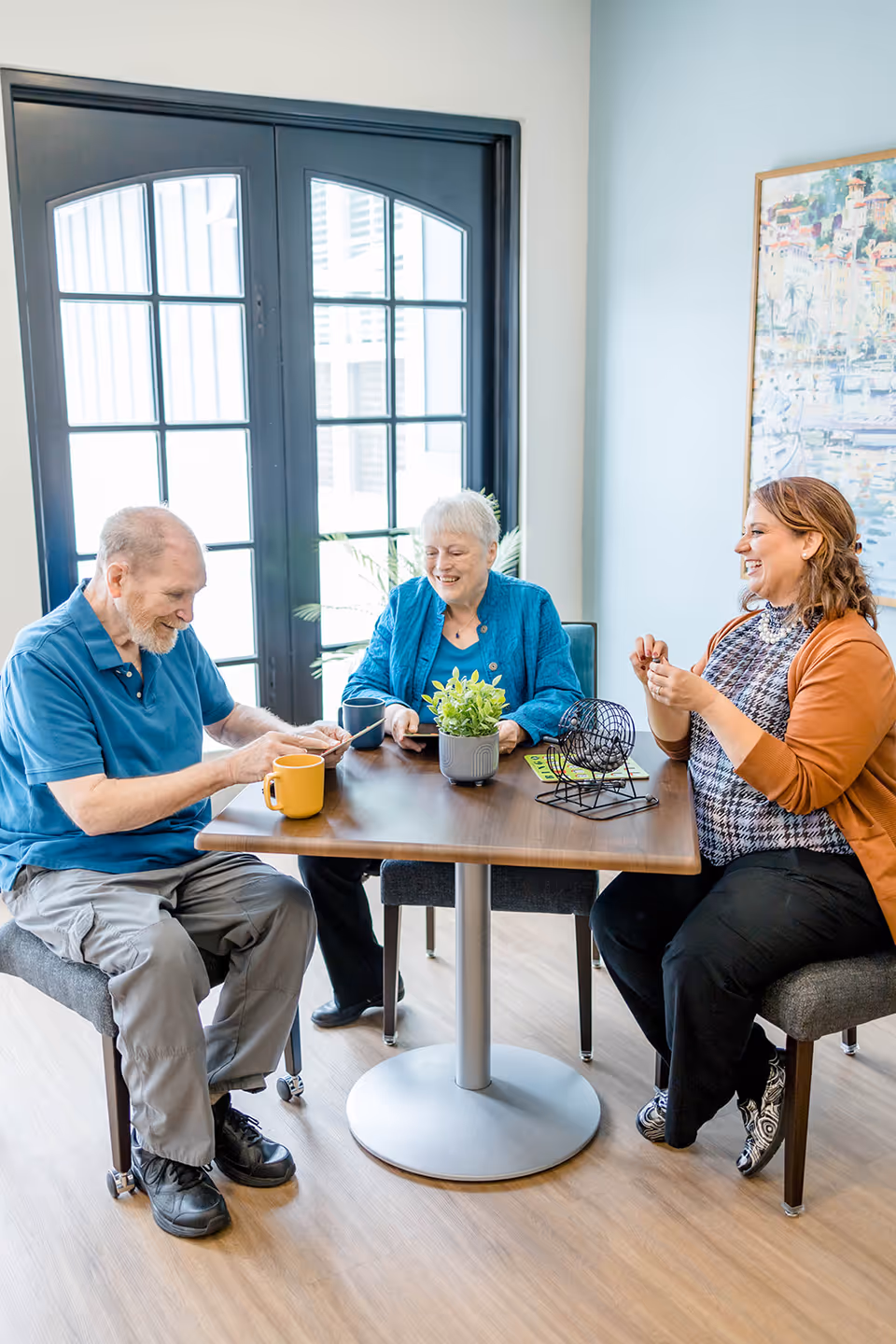 Three people sitting around a wooden table playing a game with bingo cards and a bingo cage. The group includes an elderly man, an elderly woman, and a younger woman. The room has large black-framed windows and light-colored walls with a painting on one side.