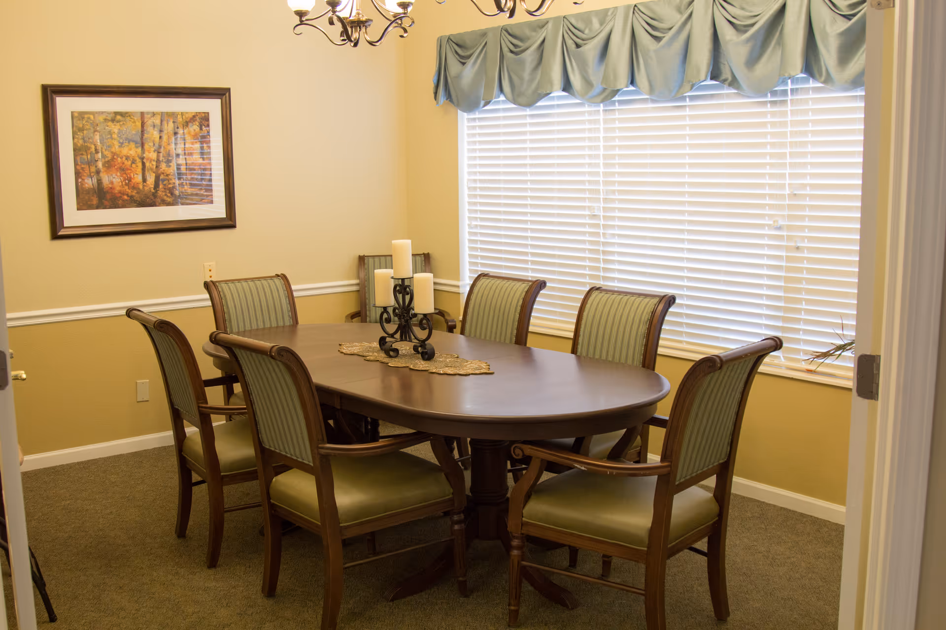 A dining room with a wooden oval table surrounded by six wooden chairs with green striped upholstery. On the table is a decorative candle holder with four white candles and a gold-colored table runner. The room has beige walls, a framed painting of autumn trees, a window with white blinds and a blue valance, and a chandelier hanging from the ceiling.