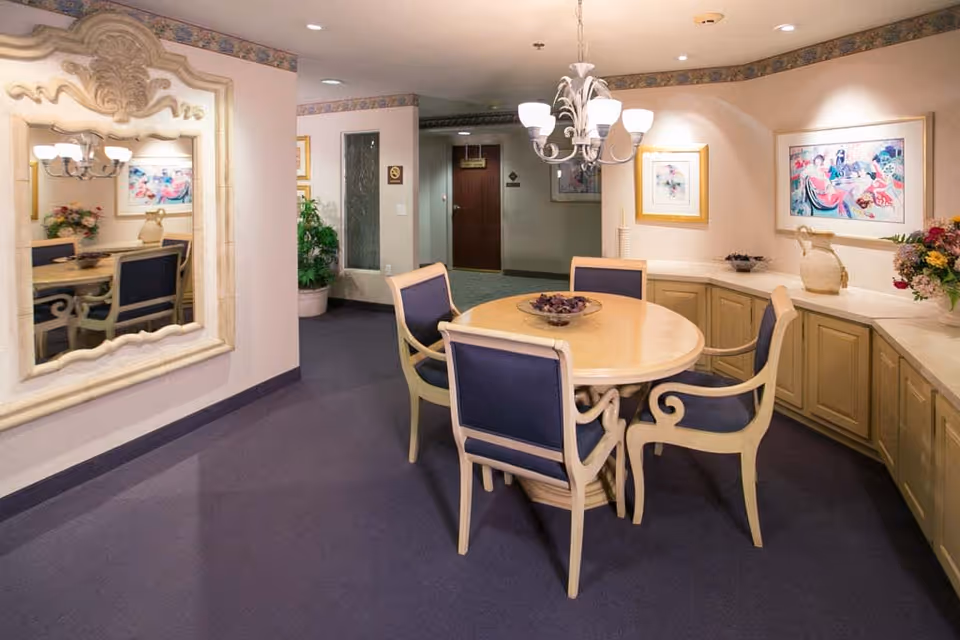 A cozy dining area with a round wooden table surrounded by four chairs with blue cushions. The room features a large ornate mirror on one wall, framed artwork, a chandelier with multiple lights hanging above the table, and built-in cabinetry along the walls with decorative items and flowers.