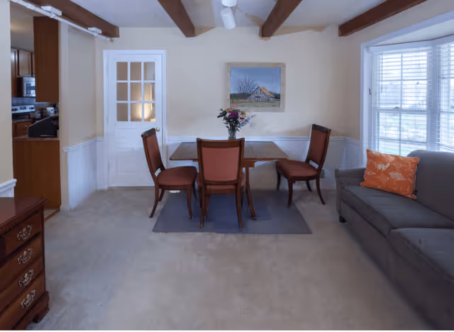 A dining area with a wooden table and four chairs, a vase with flowers on the table, a painting on the wall, a gray couch with an orange pillow, and a window with blinds letting in natural light. The room has beige walls, exposed wooden ceiling beams, and a door leading to a kitchen area.