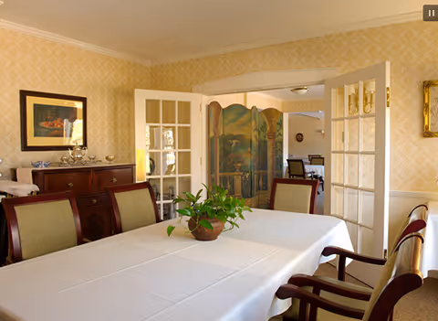 A dining room with a long table covered with a white tablecloth and surrounded by wooden chairs with green cushions. There is a potted plant centerpiece on the table. The room has yellow patterned wallpaper, a wooden sideboard with decorative items, and French doors leading to another room with additional seating and a decorative folding screen.