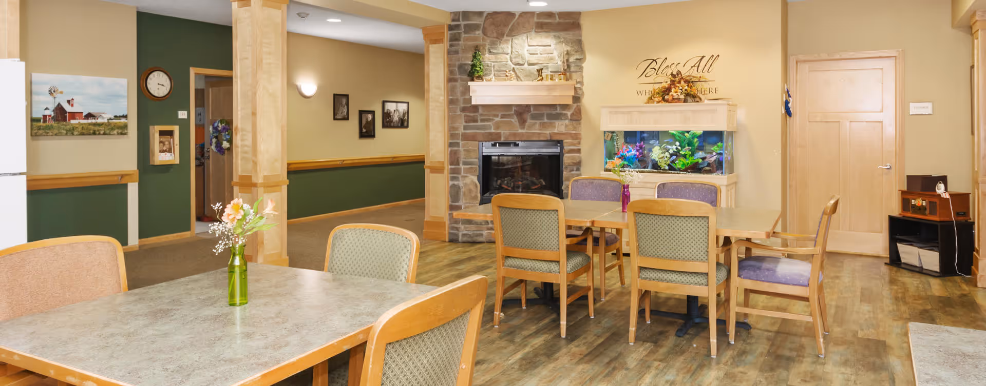 A cozy common area in an assisted living facility featuring several tables and chairs, a stone fireplace, a large fish tank, and wall decorations including framed pictures and a sign that reads 'Bless All Who Enter Here'. The room has wooden flooring and a warm color scheme with green and beige walls.