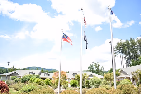 Outdoor view of a senior living facility with multiple flagpoles displaying flags, surrounded by landscaped bushes and trees, with buildings and a partly cloudy sky in the background.