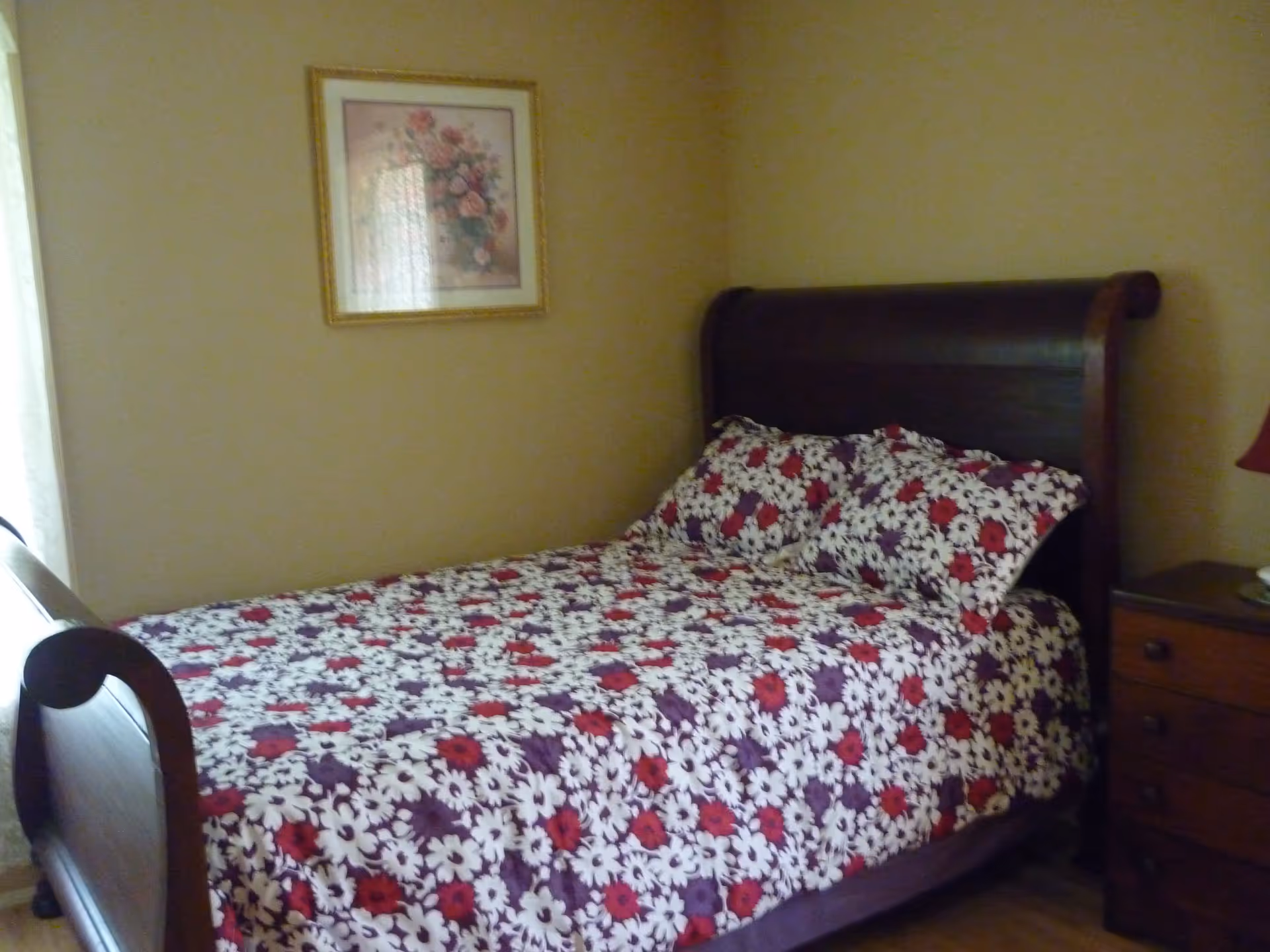 A bedroom with a wooden sleigh bed featuring a floral bedspread in red, white, and purple colors. There is a wooden nightstand with a lamp on the right side of the bed and a framed floral painting hanging on the beige wall above the bed.